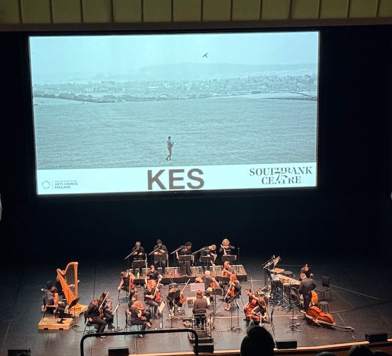 A screen showing a poster for Kes the film, beneath it is an orchestra