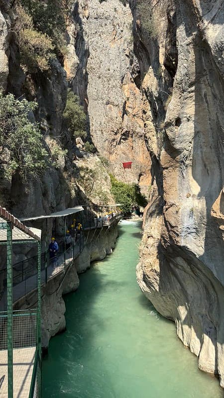 Saklikent Gorge with a blue river at the bottom on the right hand side is a covered walkway which is full of people wearing yellow safety helmets